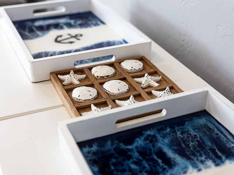 Close-up of two shallow bins with an ocean and anchor pattern at their bottom, and in the middle a wooden tic-tac-toe game that uses circular shells and starfishes